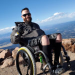 Andrew sits in his wheelchair on a mountain top, blu skies behind him
