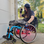 A young female with black hair wearing a black cap sitting in her neon colored wheel chair. She is doing the peace sign with her fingers.