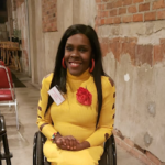 A woman with black hair. She is wearing a bright yellow dress with a red flower on it, while sitting in her wheelchair.
