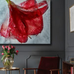 Large photograph of a red flower hanging in agray room . a golden table in front of it with a vase with flowers and a chair