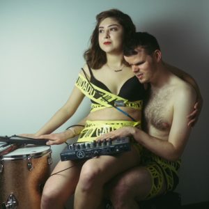 Stephanie Alma and Tommy Carroll, wearing only their underwear, seated together at a drumset. Arms and bodies intertwined, they are wrapped in caution tape. Their right hands both rest on top of a snare drum. Her left arm is draped luxuriously over his shoulder. His left arm caresses a mixing board on her lap, and his head rests on her shoulder.