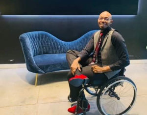 James sits in his wheelchair infront of a blue plush couch. He smiles and sits with his leg crossed. He wears a button up shirt, tie and vest with slacks and red shoes.